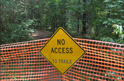 Yellow warning sign stating "No Access to Trails" is posted on an orange construction fence, with a hiking trail visible behind it.