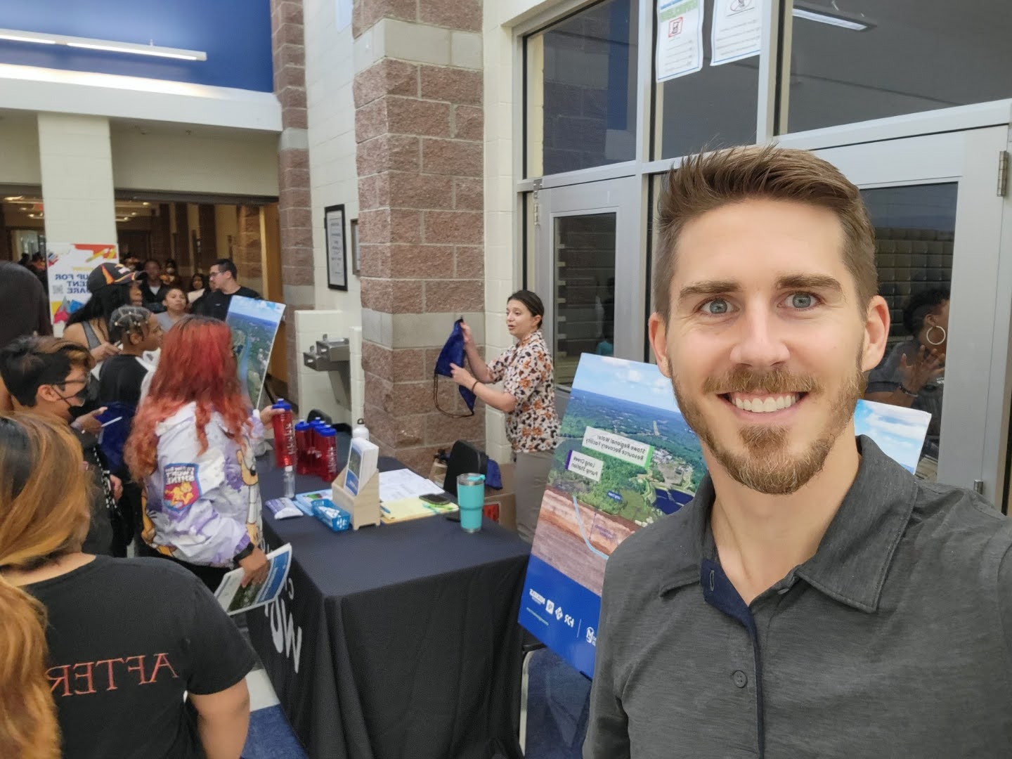 Stowe Project Manager Will Shull takes a selfie in front of Charlotte Water's information table, where a woman hands out backpacks to students.