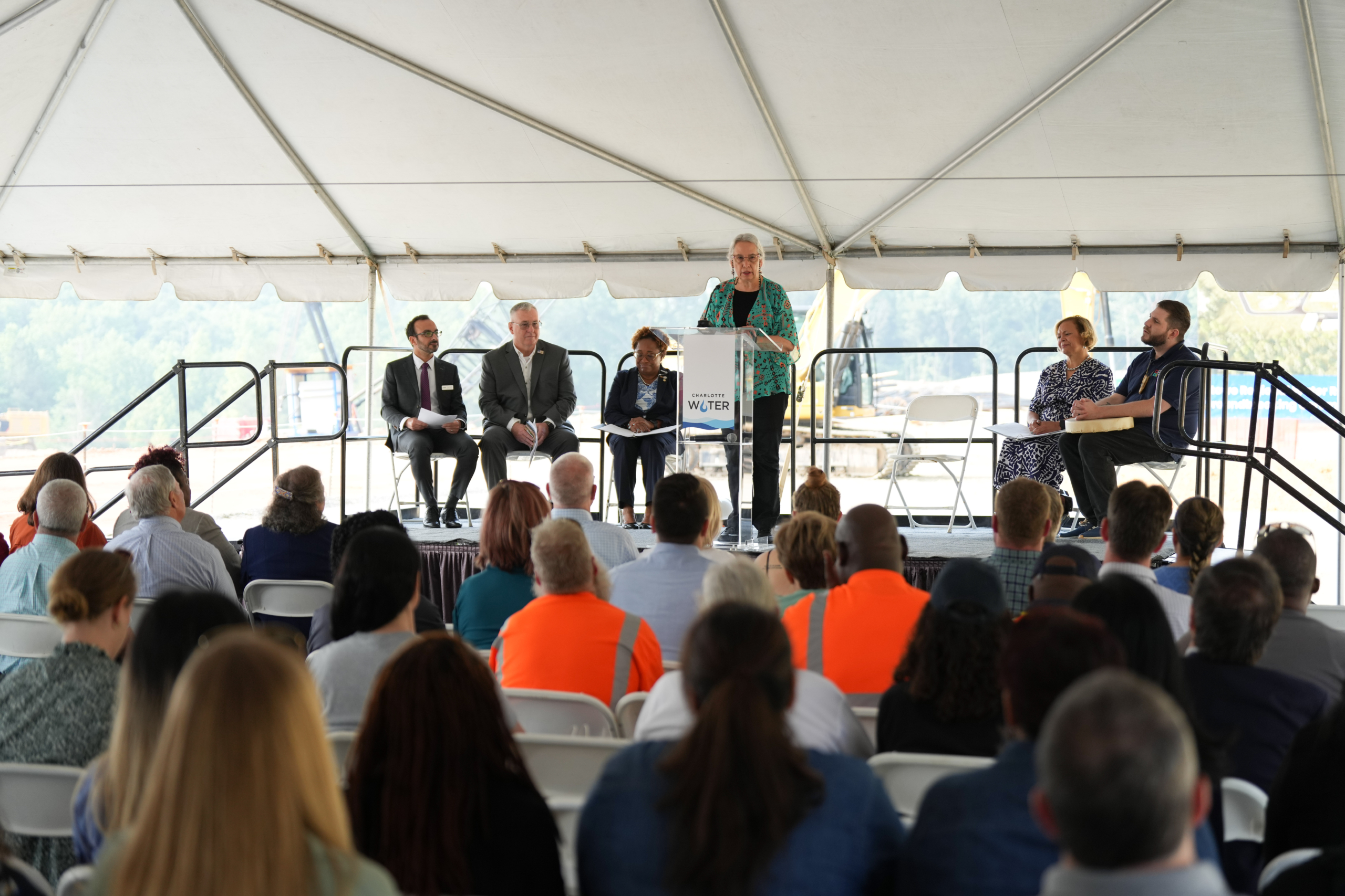 Dr. Haire speaks at the Charlotte Water podium with the other speakers watching from their seats on stage and seated guests visible in the foreground.