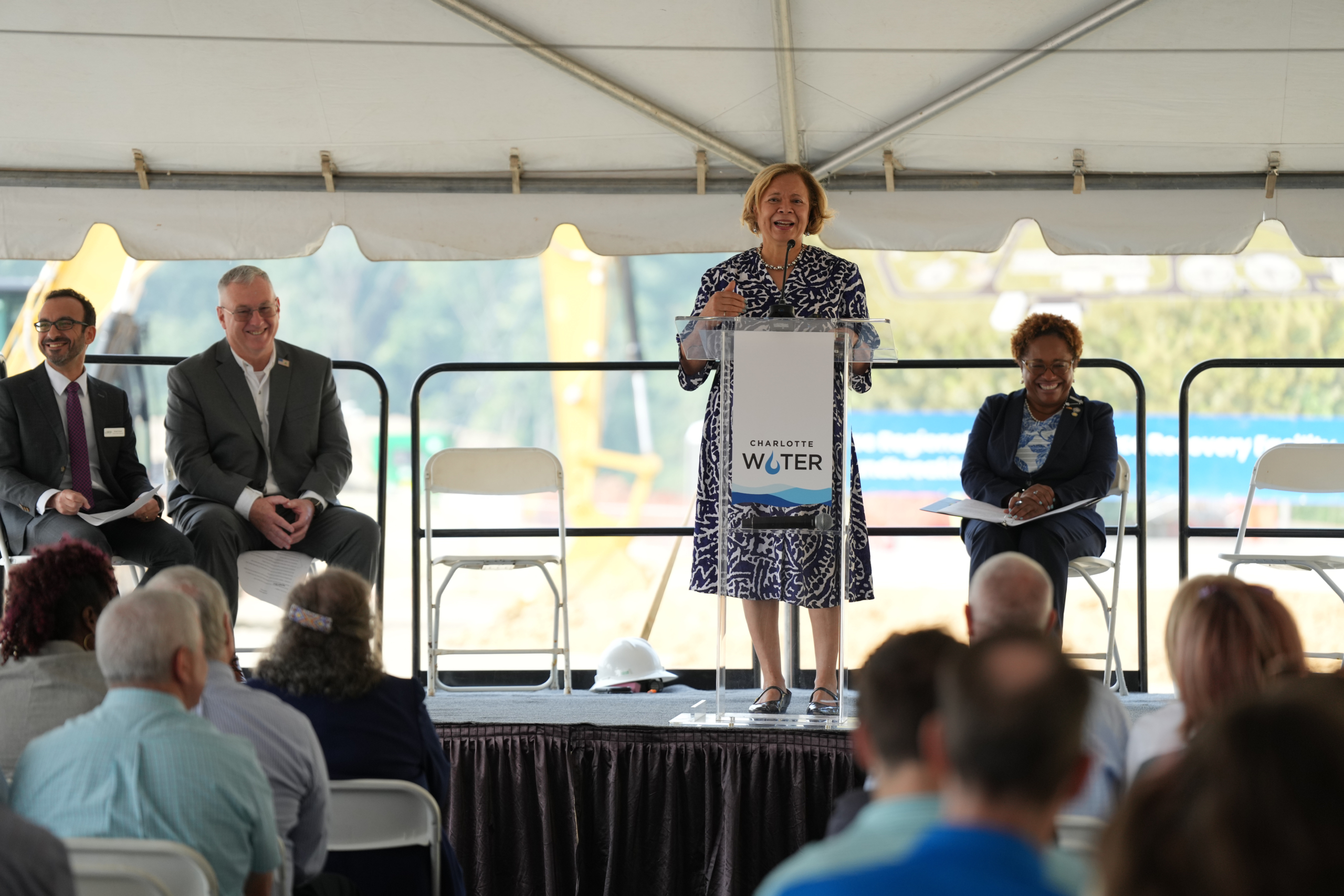 Mayor Lyles speaks at the Charlotte Water podium with the other speakers watching from their seats on stage and seated guests visible in the foreground.