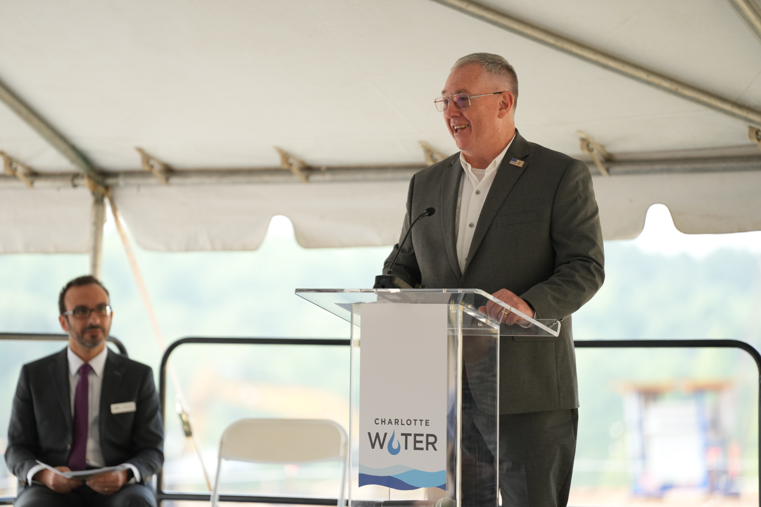 John Nicholson speaks at the Charlotte Water podium with Shadi Eskaf watching from his seat on stage behind him.