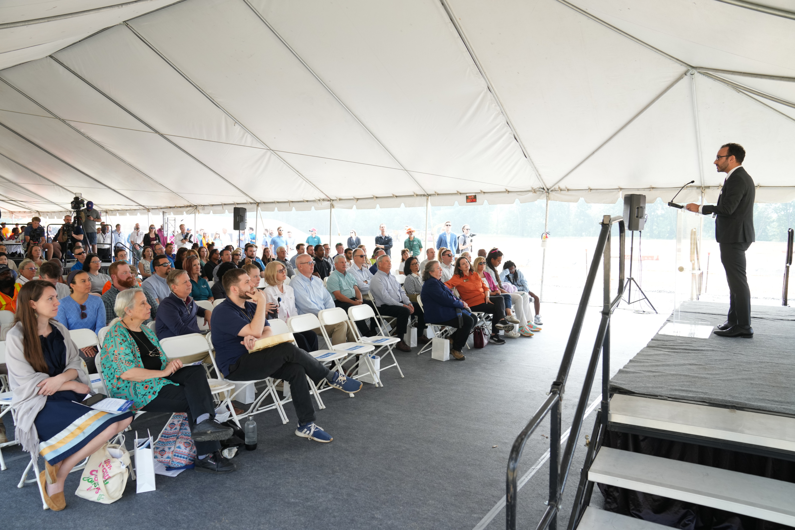 Shadi Eskaf stands at the podium on stage. Dr.  Haire and Delesslin Roo George-Warren are seen sitting in the front row with a crowd of attendees.