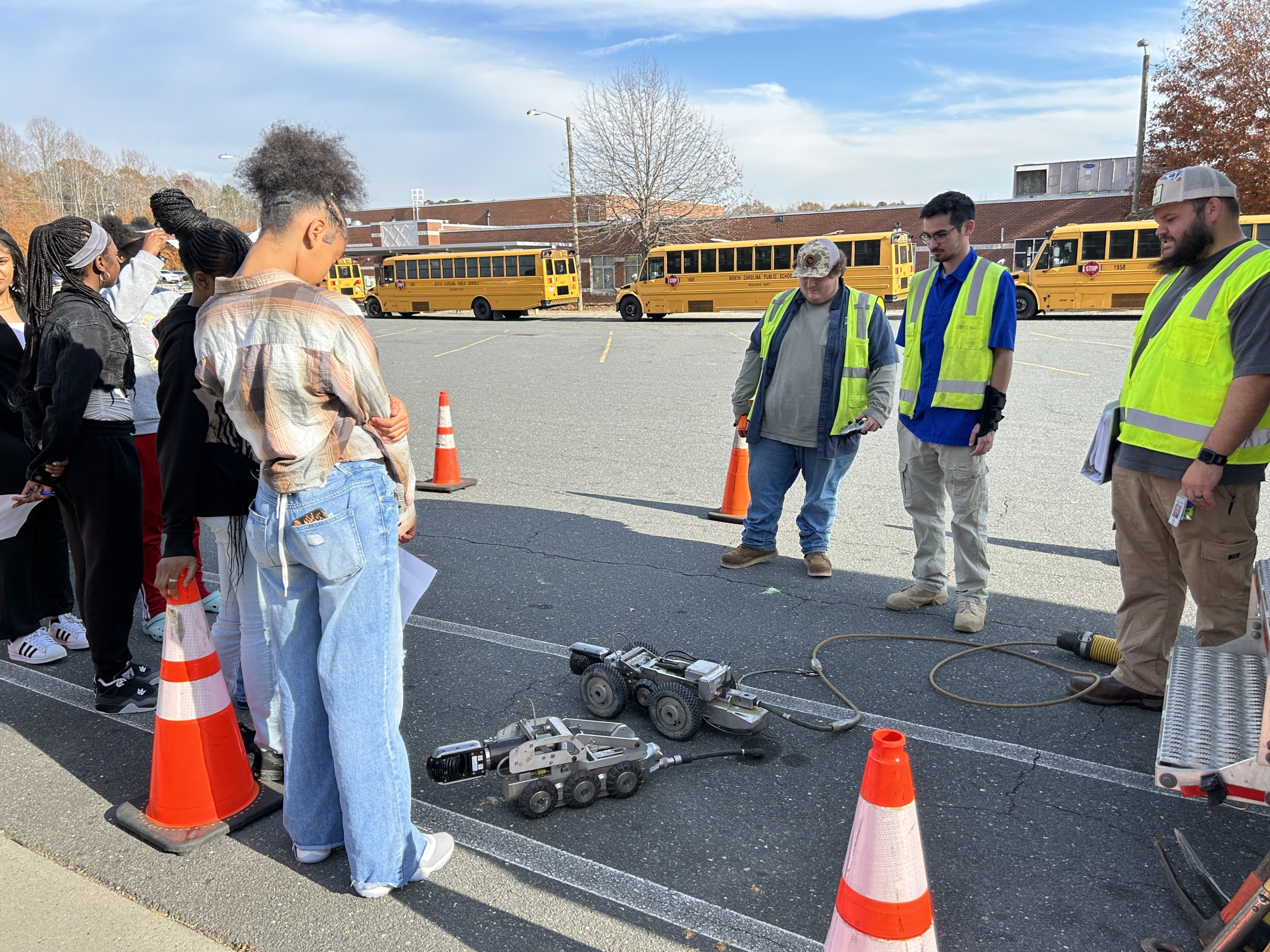 Three men in safety vests stand in a school parking lot in front of buses with a group of students. They all look down at two rubber-wheeled robots with cameras on the front of the machines. 