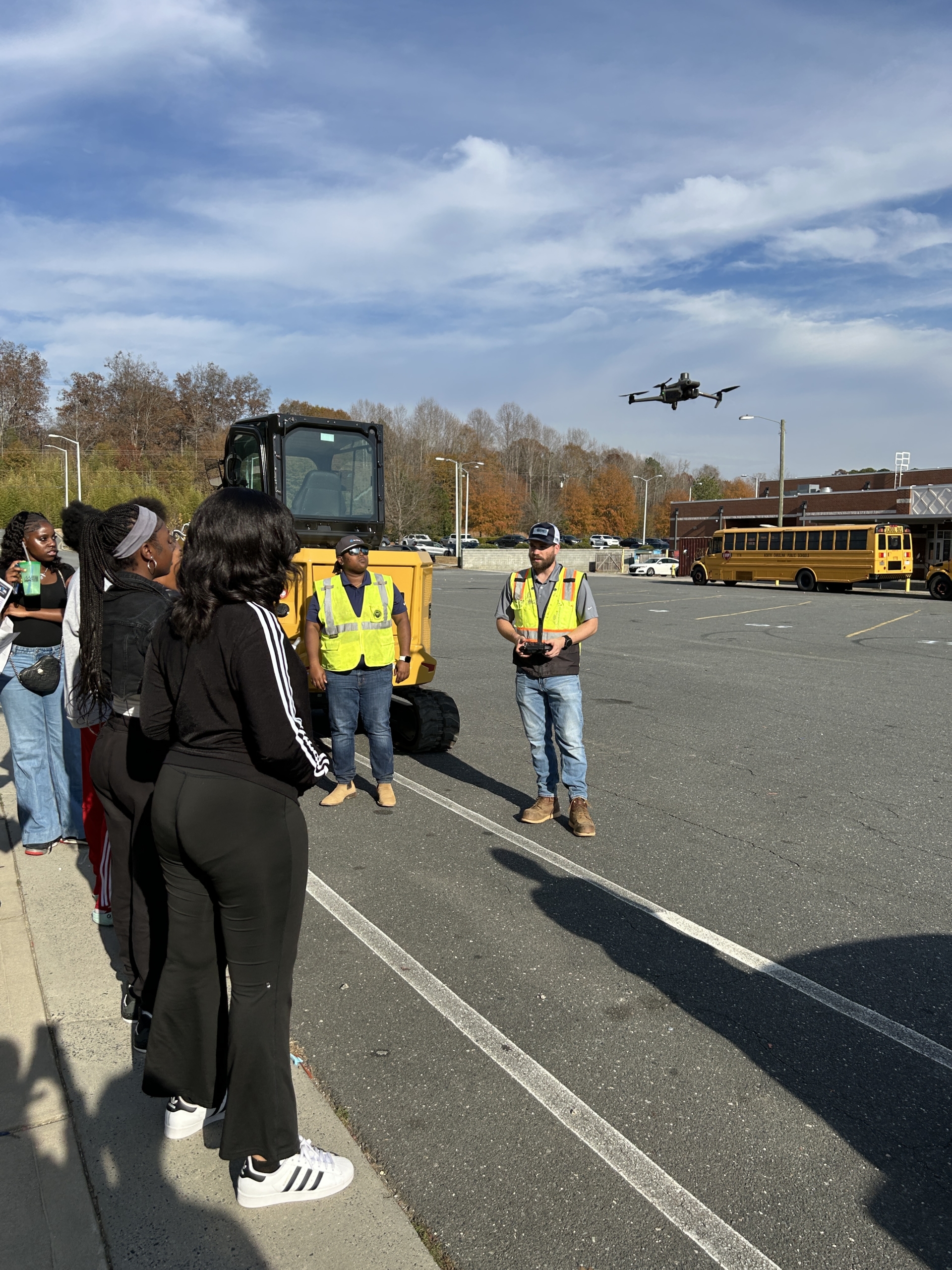 Two Garney Construction workers stand in front of a group of students in the school parking lot next to a CAT tracked vehicle with a drone hovering in the air.