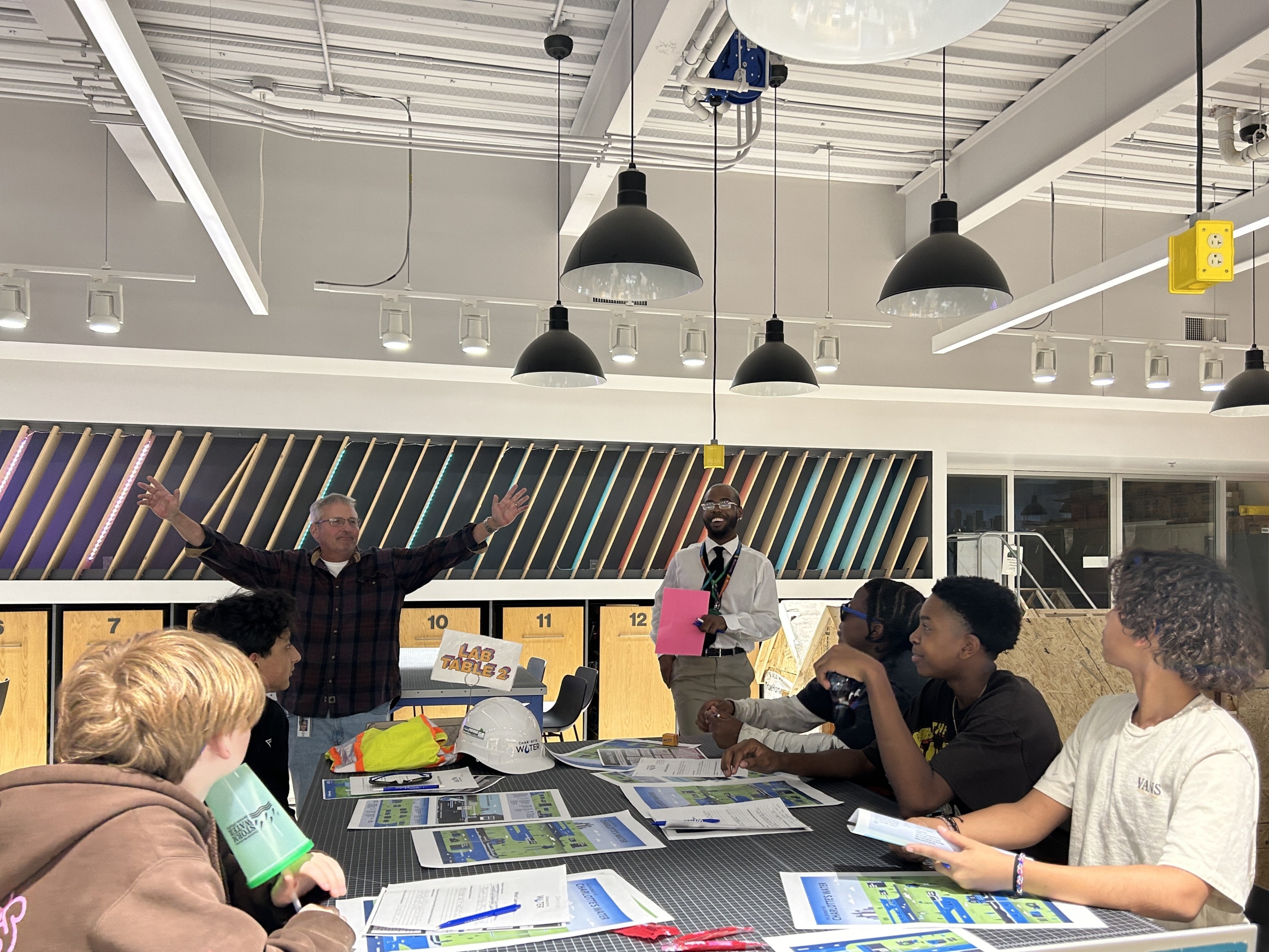 A Charlotte Water Employee stands with arms outstretched in front of a table of students with Mr. Rutley smiling at his side.