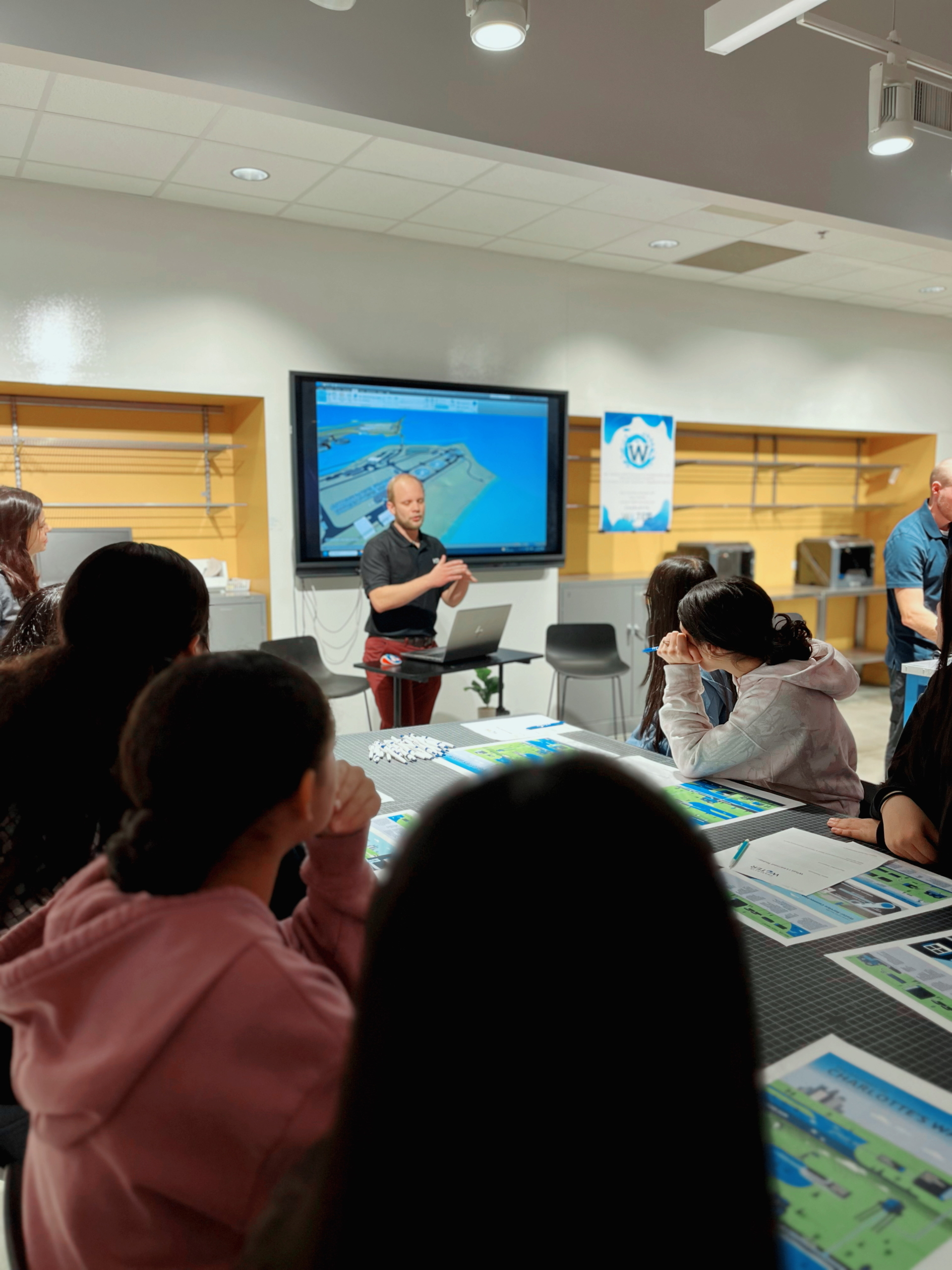 An engineer stands in front of a TV monitor displaying a 3D model of the wastewater treatment plant site as students sitting around the table look on. 