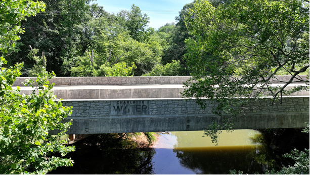 A concrete bridge with Charlotte Water's logo and fieldstone motif spans over a creek amid spring leaves.