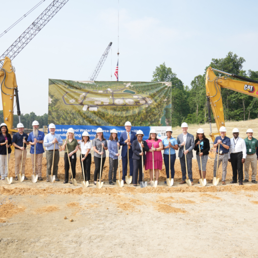 Charlotte Water Employees wearing hard hats and holding golden shovels stand in front of a banner depicting the new facility rendering hung from two excavators.