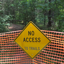 Yellow warning sign stating "No Access to Trails" is posted on an orange construction fence, with a hiking trail visible behind it.