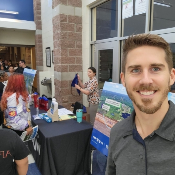 Stowe Project Manager Will Shull takes a selfie in front of Charlotte Water's information table, where a woman hands out backpacks to students.